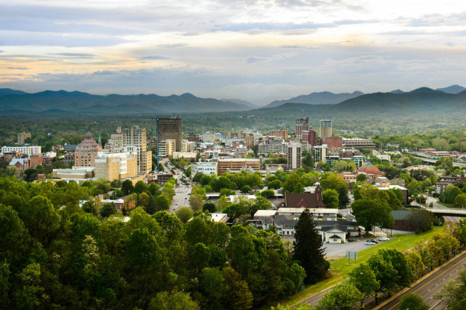 The skyline of downtown Asheville, North Carolina at sunset with green trees in the foreground and mountains in the distant background